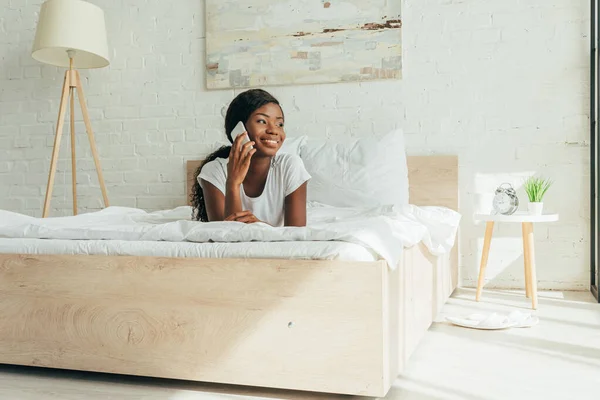 Chica afroamericana feliz acostado en la cama, hablando en el teléfono inteligente y mirando hacia otro lado - foto de stock