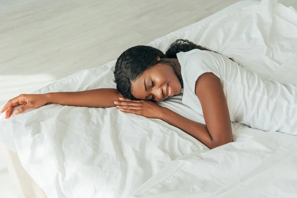Vista de ángulo alto de hermosa, sonriente chica afroamericana acostada sobre ropa de cama blanca con los ojos cerrados - foto de stock