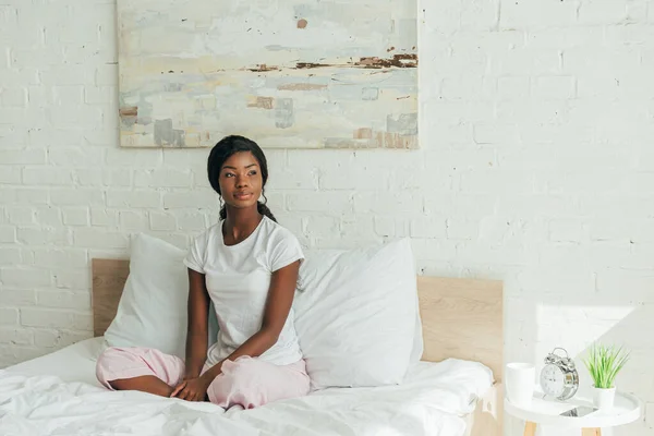 Chica afroamericana pensativa en pijama sentada en la cama con las piernas cruzadas y mirando hacia otro lado - foto de stock