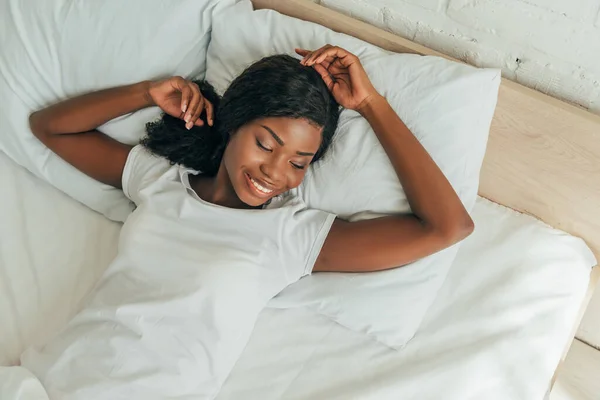 Top view of happy, attractive african american girl smiling while lying in bed — Stock Photo