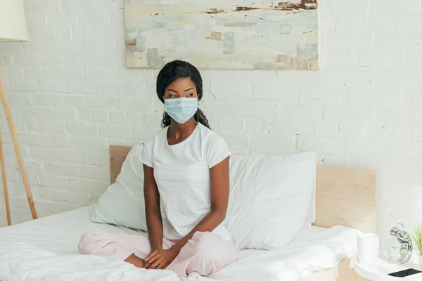 Dreamy african american woman in medical mask sitting in bed with crossed legs and looking away — Stock Photo