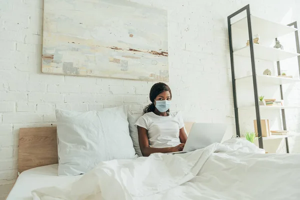 Afroamericano freelancer en máscara médica sentado en la cama con el ordenador portátil y mirando a la cámara - foto de stock