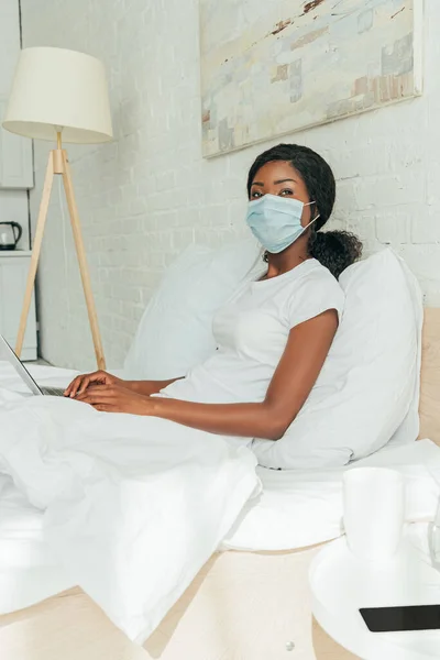 Afroamericano freelancer en máscara médica sentado en la cama con el ordenador portátil y mirando a la cámara - foto de stock