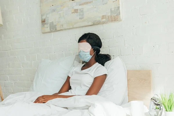 Jovem afro-americana em máscara médica e máscara de sono descansando enquanto sentado na cama — Fotografia de Stock