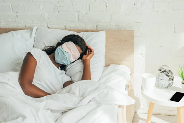African american woman in sleep mask and medical mask sleeping on white bedding — Stock Photo