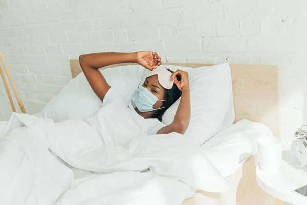 Africano americano ragazza toccare sonno maschera mentre sdraiato e letto e guardando fotocamera — Foto stock