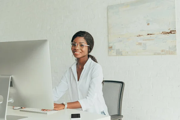 Cheerful african american freelancer smiling at camera near computer monitor at home — Stock Photo
