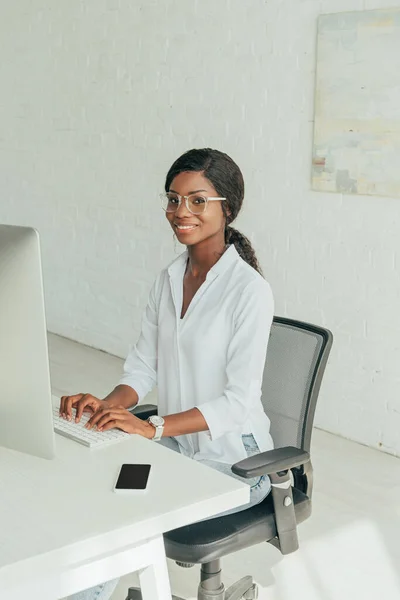 Feliz afroamericano freelancer escribiendo en el teclado mientras sonríe a la cámara - foto de stock