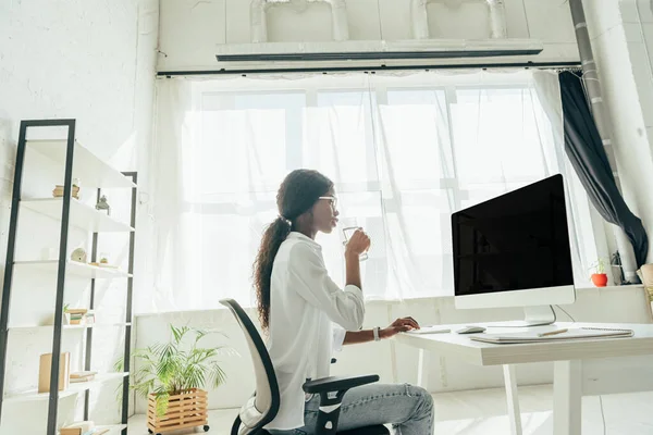 Vista lateral del freelancer afroamericano sosteniendo un vaso de agua y mirando el monitor de computadora con pantalla en blanco mientras trabaja en casa - foto de stock