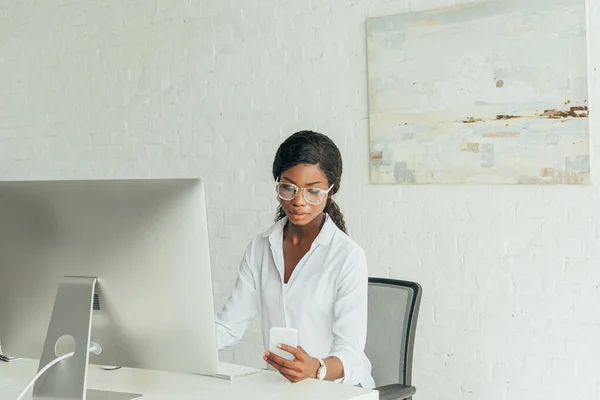 Grave afroamericano freelancer charlando en el teléfono inteligente cerca del monitor de la computadora en casa - foto de stock