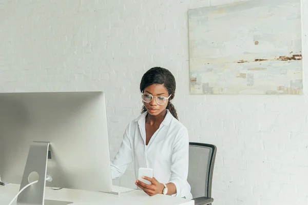 Serious african american freelancer chatting on smartphone near computer monitor at home — Stock Photo