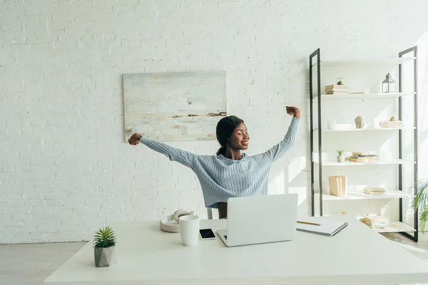 Fröhliche afrikanisch-amerikanische Freelancerin dehnt sich mit geschlossenen Augen, während sie am Tisch neben dem Laptop sitzt — Stockfoto