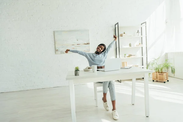 Lächelnde afrikanisch-amerikanische Freelancerin, die sich dehnt, während sie neben dem Laptop im geräumigen Wohnzimmer sitzt — Stockfoto