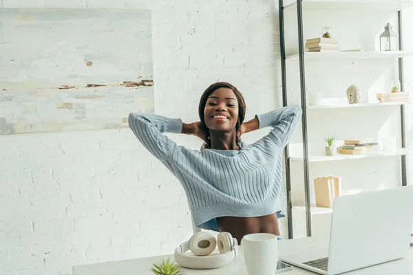 Freelancer afro-americano alegre alongamento enquanto sentado perto de laptop, fones de ouvido sem fio e xícara de café — Fotografia de Stock
