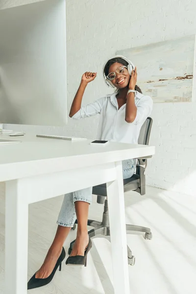 Foco seletivo de sorrir freelancer afro-americano em fones de ouvido sem fio ouvir música perto do monitor do computador — Fotografia de Stock