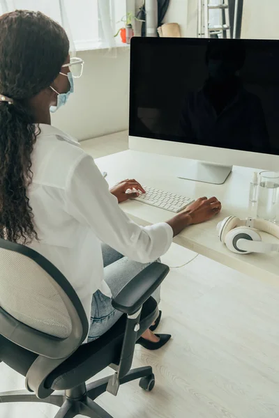 Jovem freelancer afro-americano em máscara médica olhando para monitor de computador com tela em branco — Fotografia de Stock
