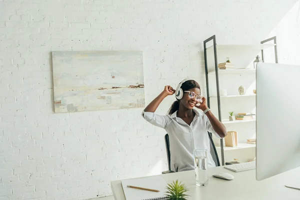 Cheerful african american freelancer listening music in wireless headphones near computer monitor at home — Stock Photo