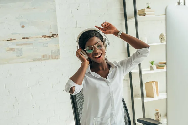 Happy african american freelancer gesturing and touching wireless headphones while listening music — Stock Photo