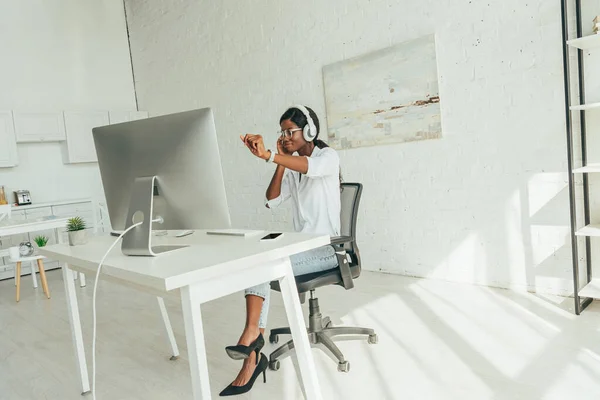 Cheerful african american freelancer gesturing while listening music in wireless headphones near computer monitor — Stock Photo