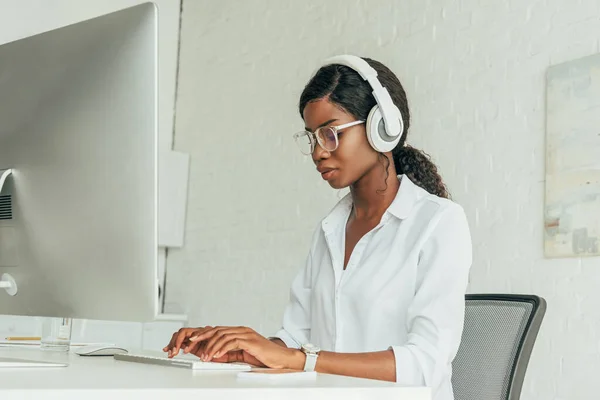 Attentive african american freelancer in wireless headphones and glasses typing on computer keyboard — Stock Photo