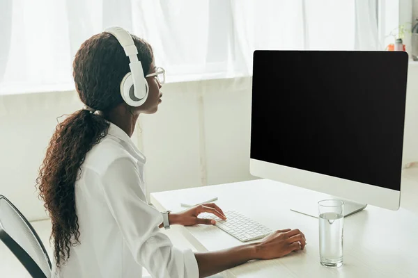 Young african american freelancer in wireless headphones looking at computer monitor with blank screen — Stock Photo