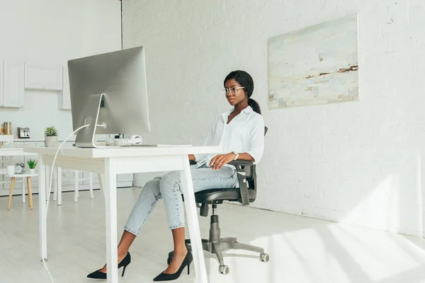 Joven afroamericano freelancer en blusa blanca, jeans y zapatos de tacón alto mirando el monitor de computadora en casa - foto de stock