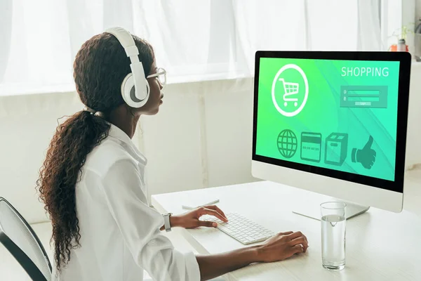 Young african american freelancer in wireless headphones looking at computer monitor with shopping website — Stock Photo