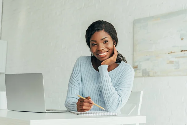 Fröhliche afrikanisch-amerikanische freie Mitarbeiterin lächelt in die Kamera, während sie neben Laptop sitzt — Stockfoto