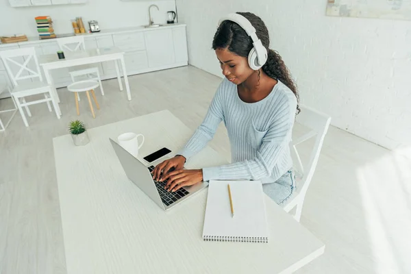 Joven afroamericano freelancer en auriculares inalámbricos trabajando en el ordenador portátil en la cocina - foto de stock