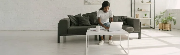 Горизонтальное изображение африканского фрилансера, работающего на ноутбуке в просторной гостиной — стоковое фото