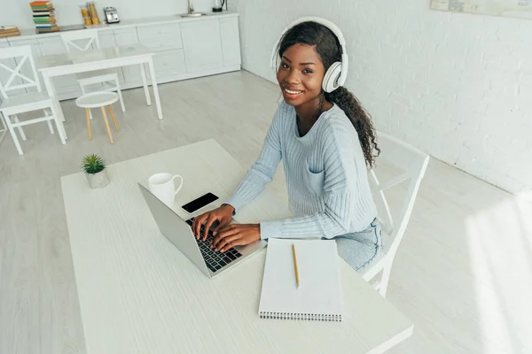 Vista de ángulo alto de sonriente freelancer afroamericano en auriculares inalámbricos que trabajan en el ordenador portátil en la cocina - foto de stock