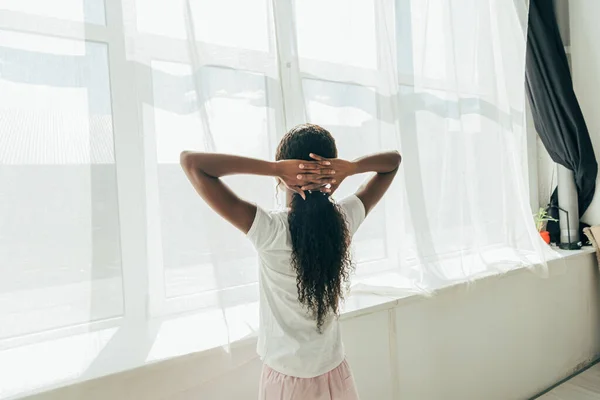 Vista trasera de chica afroamericana de pie junto a la ventana bajo el sol con las manos detrás de la cabeza - foto de stock