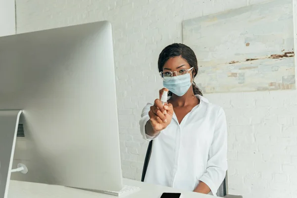 Молодой африканский американский фрилансер в медицинской маске показывает антисептический спрей, сидя рядом с монитором компьютера дома — стоковое фото