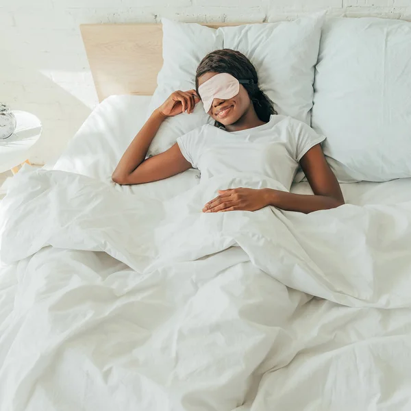 Visão de alto ângulo da menina americana africana em máscara de sono sorrindo enquanto deitada na cama — Fotografia de Stock