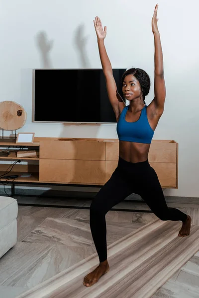 African american sportswoman standing in high lunge pose with hands in air and smiling on yoga mat in living room — Stock Photo