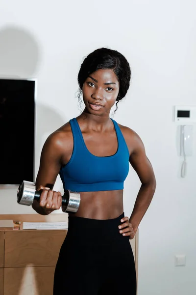 Desportista afro-americana com a mão no quadril treinando com halteres e olhando para a câmera na sala de estar — Fotografia de Stock