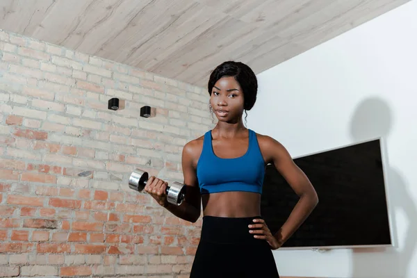 Afroamerikanische Sportlerin mit Hand auf Hüfttraining mit Hantel und Blick in die Kamera im Wohnzimmer — Stockfoto