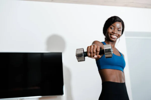 African american sportswoman holding dumbbell with outstretched hand, smiling and looking at camera in living room — Stock Photo