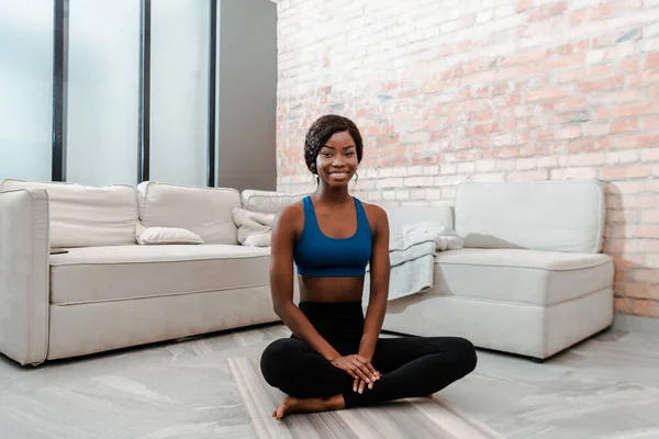 Afroamerikanische Sportlerin lächelt in die Kamera und sitzt in Lotus-Pose auf Yogamatte im Wohnzimmer — Stockfoto