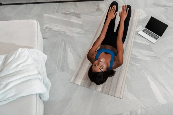 Vista aérea de la deportista afroamericana practicando yoga en el piso cerca de la computadora portátil en la sala de estar - foto de stock