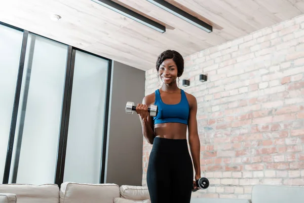 Femme sportive afro-américaine souriante, s'entraînant avec des haltères et regardant la caméra dans le salon — Photo de stock