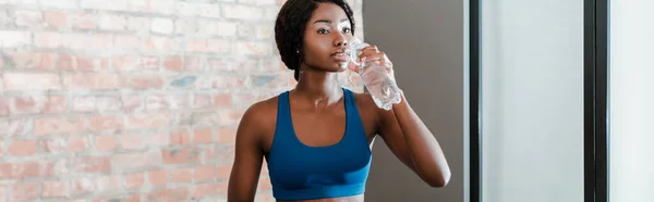 Panoramaorientierung einer afrikanisch-amerikanischen Sportlerin, die Wasser im Wohnzimmer trinkt — Stockfoto