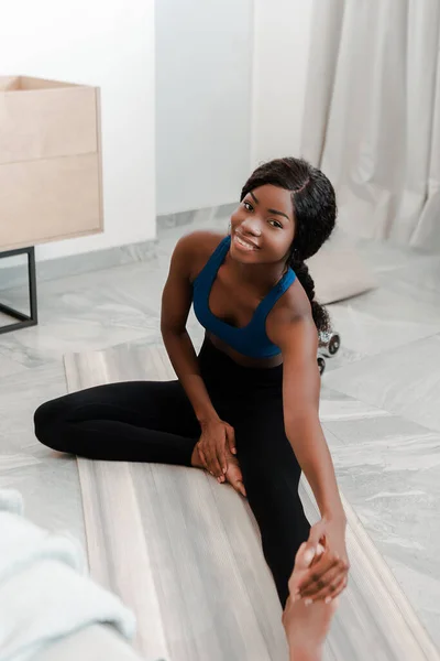 Deportista afroamericana estirándose, sonriendo y mirando la cámara en la esterilla de yoga en la sala de estar - foto de stock