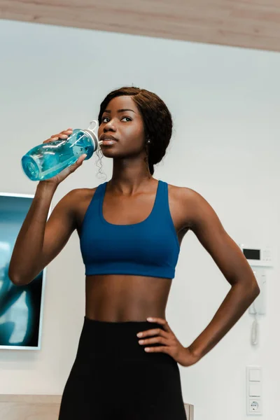 African american sportswoman with hand on hip drinking water and looking away in living room — Stock Photo