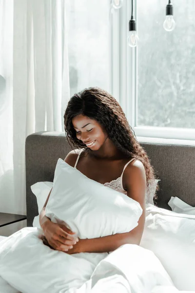 African american woman holding pillow and smiling on bed in bedroom — Stock Photo