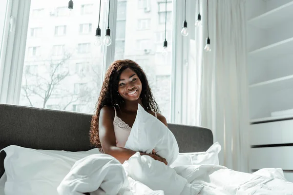 Donna afroamericana che tiene il cuscino, sorride e guarda la macchina fotografica sul letto in camera da letto — Foto stock