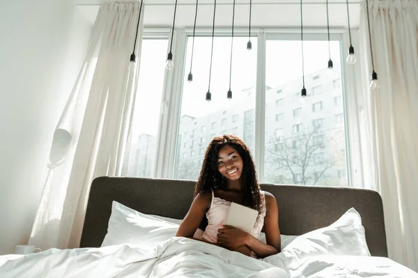 Donna afroamericana con libro sorridente e guardando la macchina fotografica sul letto in camera da letto — Foto stock