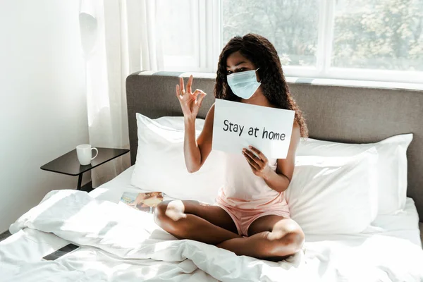 African american woman in medical mask with crossed legs showing okay sign and placard with stay at home lettering on bed in bedroom — Stock Photo