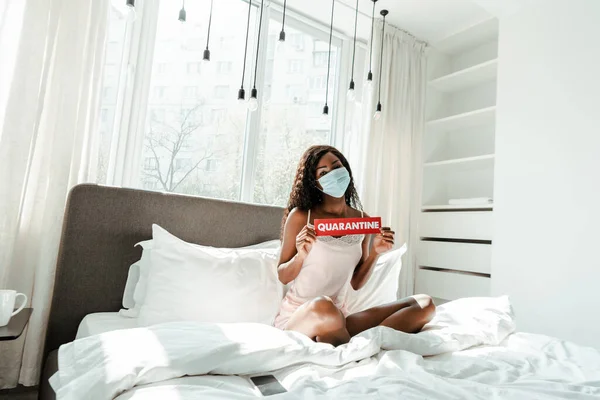 African american woman in medical mask with crossed legs showing card with quarantine lettering on bed in bedroom — Stock Photo