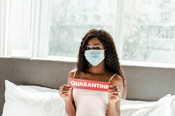 Chica afroamericana en máscara médica mostrando tarjeta con letras de cuarentena en la cama en el dormitorio - foto de stock
